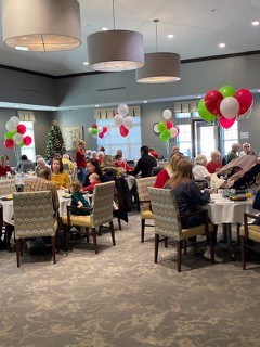 Groups of people celebrating Christmas sitting around tables with red, and green, balloons in the dining room. Grey walls and brown patterned carpet