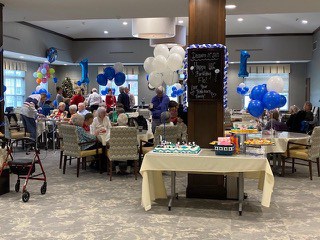 A party with a group of people in the background. Blue and white balloons and tables with yellow table cloths.