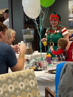 A family sitting around a table with White and Green balloons in a dining room. A baby is looking at a server holding orange juice and wearing an elf costume, and the family is laughing.