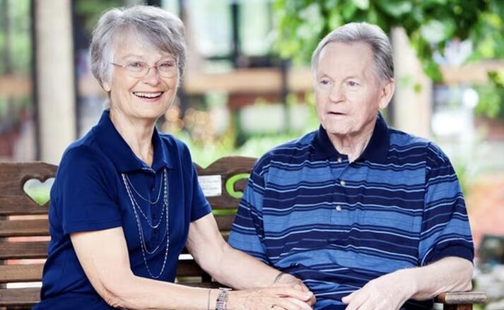 Elderly couple sit together on a park bench, holding hands. They are both happy and enjoying the sunshine. Traditions at Solana.