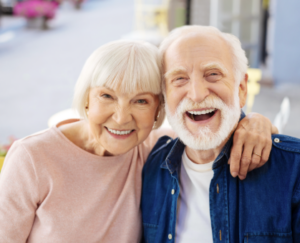 An older couple posing together outside; the woman rests her hand on the man’s shoulder, and both are smiling.