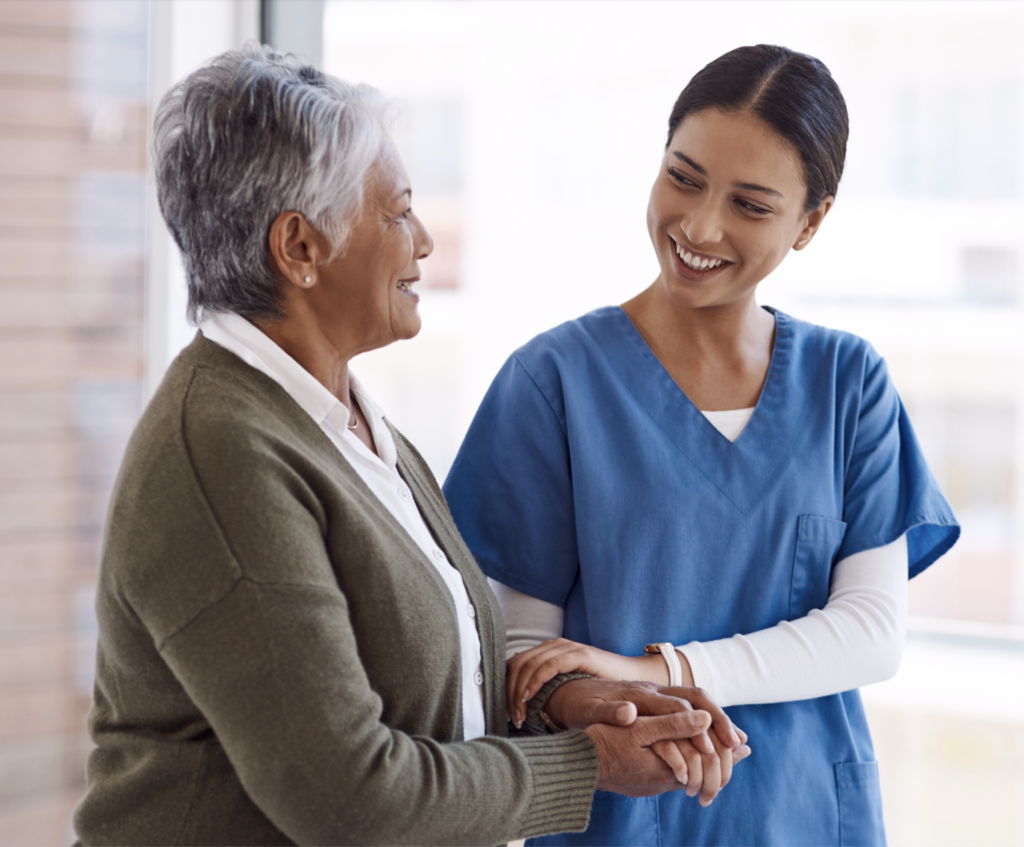A caregiver in blue scrubs holding a senior woman with short gray hair down a hall with tall windows.